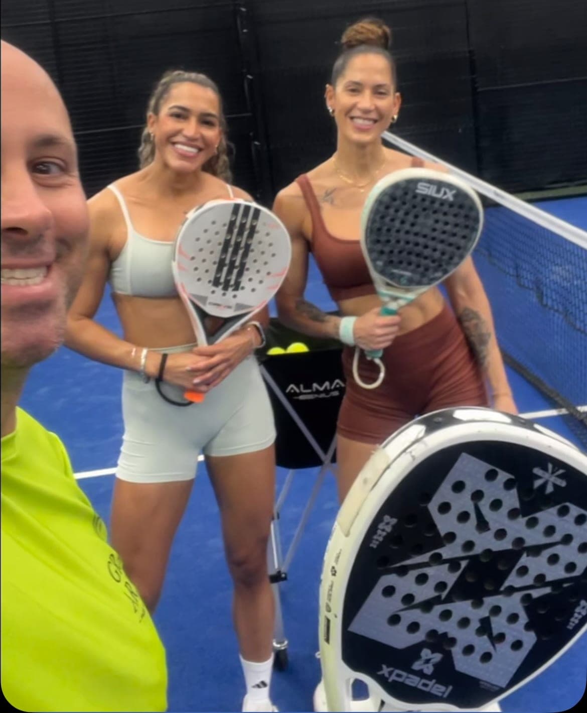 Professor e duas alunas sorrindo com as raquetes na quadra de padel, antes da aula